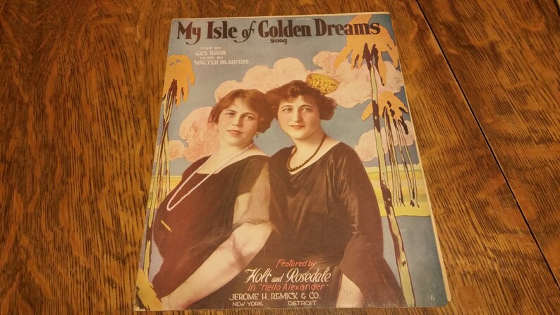 May include: A vintage sheet music cover for the song "My Isle of Golden Dreams" featuring two women in black dresses. The cover features a tropical scene with palm trees and clouds.