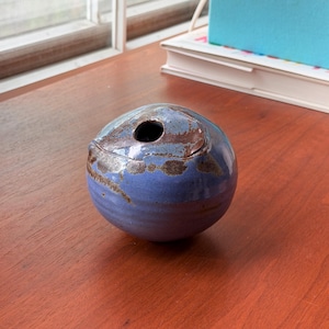 Könnte beinhalten: Kleine, runde Keramikvase mit blauer und brauner Glasur. Die Vase hat eine einzelne Öffnung an der Oberseite und steht auf einer Holzoberfläche. Die Farbpalette umfasst Blau-, Braun- und Weißtöne.