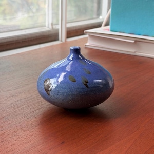 May include: A small, round, blue ceramic vase with a narrow neck. The vase has a speckled design with dark brown spots and a gradient from a lighter blue at the base to a darker blue at the top. The vase is sitting on a wooden surface.