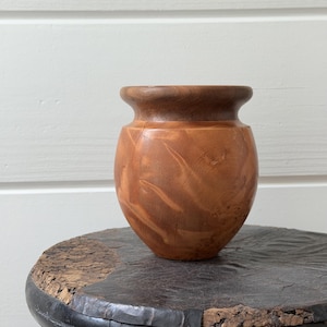 May include: A small, handcrafted wooden vase with a rounded body and a flared rim. The vase is made of light brown wood with darker brown accents, showcasing the natural grain patterns. It sits on a dark, textured surface.