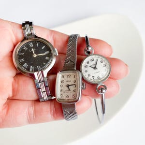 May include: A selection of vintage wristwatches with silver metal straps and watch faces. One watch has a black face with Roman numerals, another has a rectangular face with the word "ЧАЙКА". The third watch has a round face.