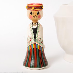 May include: A wooden doll with a painted face, wearing a white, green, red, and blue dress with a red and yellow headband. The doll is standing on a white surface.