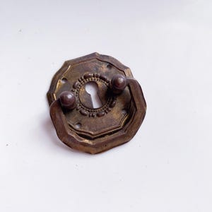 Op de afbeelding: Antieke messingkleurige ladeknop met sleutelgat. De knop heeft een achthoekige achterplaat met een centraal sleutelgat en een kleine handgreep. De hardware heeft een vintage uitstraling.