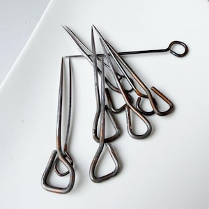 May include: A collection of six dark, metallic, pointed sewing pins with looped, triangular-shaped heads. The pins feature a weathered, oxidized finish with visible copper and charcoal tones, arranged in a cluster on a smooth, white surface.