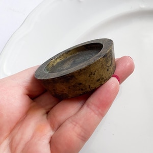 May include: A small, antique brass ashtray with a circular, shallow bowl. The ashtray has a dark, aged patina, showing signs of wear. The ashtray is held in a hand, with a white plate in the background.