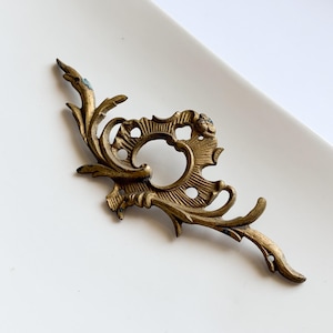 Könnte beinhalten: Ein längliches, antikes Schlüsselschild aus Messing mit detaillierten, symmetrischen Schnörkeln und blattartigen Motiven. Der Metallbeschlag weist eine verwitterte, dunkle Patina auf und besitzt eine zentrale Öffnung sowie zwei Montagelöcher.
