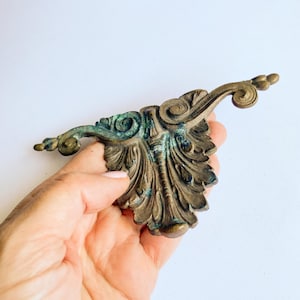 Può includere: Elemento di arredo antico in ottone con un elaborato design a foglie e volute. L'ottone presenta una patina invecchiata con sfumature verdi e marroni, che ne esaltano il fascino vintage. Ideale per progetti di restauro.