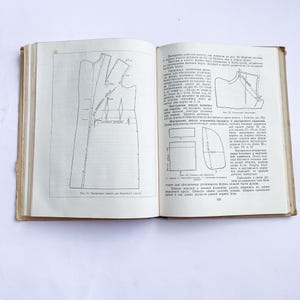 Peut inclure: Un livre ancien ouvert, avec des diagrammes et du texte détaillés, probablement pour des patrons de couture. Les pages sont vieillies, montrant de l'usure. Les diagrammes présentent des contours de pièces de vêtements, avec des mesures et des annotations. Le livre est ouvert sur une surface blanche.