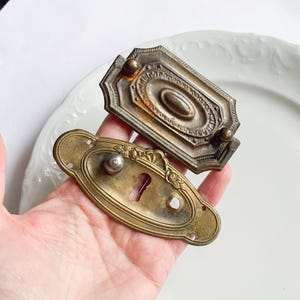 May include: Two antique brass drawer pulls and escutcheons. One is a rectangular pull with an oval center and a decorative border. The other is an oval escutcheon with a keyhole and floral accents. Both have a vintage, aged appearance.