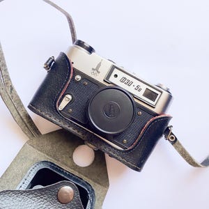 May include: Vintage silver and black camera with a black lens cap and leather case. The camera has the text "Зоркий-58" and a logo on the top. The camera is in a black leather case with a gray strap.