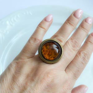 Puede incluir: Un gran anillo de cóctel redondo con una banda de metal color bronce. El anillo presenta una piedra grande de color ámbar, tallada en cabujón. El anillo se lleva en un dedo, mostrando el tamaño y el color de la joya.