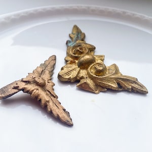 Könnte beinhalten: Zwei goldfarbene, dekorative Metallstücke mit filigranen Blumendesigns. Ein Stück ist ein Eckdesign mit einem Blumenmotiv, während das andere ein kleineres, eckigeres Stück ist.