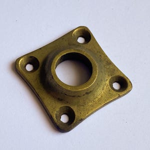 May include: A vintage brass fitting with a diamond-shaped base and a central circular aperture. Four evenly spaced holes are located at each corner of the base. The brass has a slightly aged, tarnished appearance.