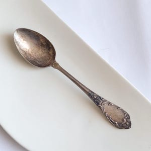 May include: A vintage silver spoon with an oval bowl and ornate handle detailing. The spoon is placed on a white rectangular plate. The spoon's surface shows signs of age and use, adding to its antique charm.