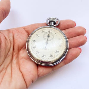 May include: A silver pocket watch with a white face and black hands. The watch has a black ring around the edge with numbers from 5 to 60. The watch face also has a smaller circle with numbers from 1 to 30. The watch has the word "Jaeger" printed on the face.
