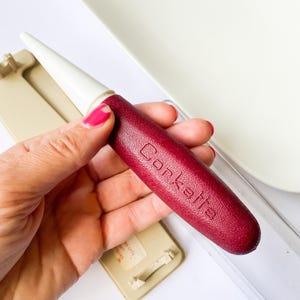 May include: A burgundy-colored cork puller with the word "Corkette" embossed on the handle. The puller has a white tip and is held near a white surface and a beige object.