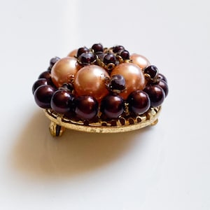 May include: A vintage brooch with faux pearls in brown and peach hues. The pearls are set on a gold-toned filigree base with small feet. A classic accessory for adding a touch of elegance to any outfit.