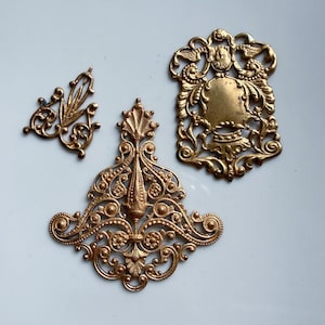 May include: Three gold-toned metal filigree ornaments featuring intricate scrollwork, floral motifs, and symmetrical patterns. The collection includes one small triangular piece, one large fan-shaped piece, and one rectangular piece with a central shield.