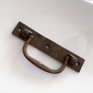 May include: A rustic, antique-style, metal drawer pull with a curved handle and two decorative knobs. The pull is a dark brown color with a worn, aged finish.