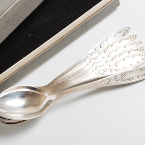 Könnte beinhalten: Ein Set aus sechs silbernen Löffeln mit verzierten Designs, gestapelt und fächerförmig angeordnet. Die Löffel befinden sich in einer grauen Schachtel mit schwarzer Auskleidung.