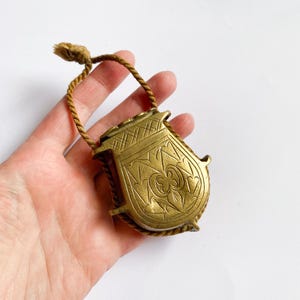 May include: A vintage brass container with intricate engraved patterns and a woven cord handle. The container has a shield-like shape and a tarnished gold finish, suggesting age and use. The cord is a light brown color.