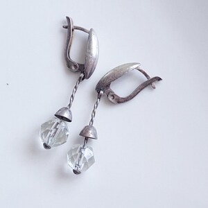 May include: A pair of silver-toned drop earrings. Each earring features a twisted wire, a small cap, and a clear, faceted crystal pendant. The earrings have a lever-back closure. The jewelry is displayed on a white surface.