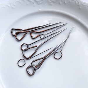 May include: A collection of antique steel and copper hat pins, arranged on a white, ornate plate. The pins vary in length and feature looped or circular head designs. The metal has a rustic, aged appearance.