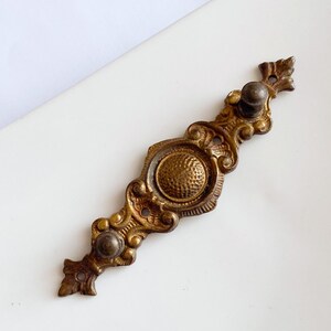 May include: Ornate antique brass drawer pull with a central textured circle and decorative scrollwork. The hardware features two small knobs and leaf-shaped accents on each end. A vintage-style cabinet or furniture accessory.