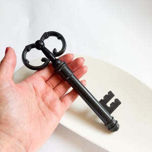 Pode incluir: Uma chave decorativa grande em preto mate com um cabo ornamentado em volutas e um eixo cilíndrico. A chave apresenta um padrão de palhetão distinto na extremidade, caracterizado por recortes retangulares e um acabamento liso e sólido.
