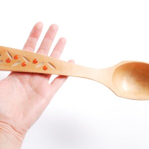 Könnte beinhalten: Ein Holzlöffel mit einem geschnitzten Design aus Blättern und roten Beeren. Der Löffel wird in einer Hand gehalten.