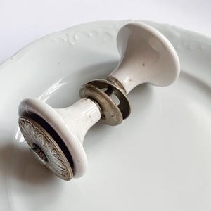 Może przedstawiać: Para antycznych białych ceramicznych gałek do drzwi z metalowymi okuciami. Gałki mają zaokrąglony kształt z dekoracyjnym kwiatowym wzorem na podstawie. Okucia obejmują metalowe podkładki i pręt łączący.