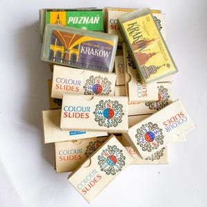 May include: A collection of vintage color slides in small cardboard boxes. The boxes feature the words "COLOUR SLIDES" in red and blue, along with images of Polish cities like Poznan and Krakow. The boxes are stacked on a white surface.