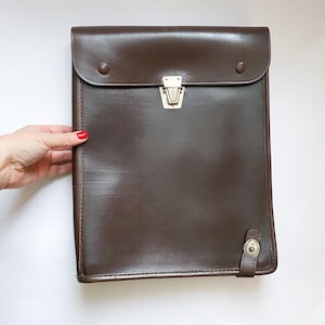 May include: A brown leather portfolio with a flap closure and a silver-colored clasp. The portfolio has a textured surface and a small strap with a button closure on the bottom. The portfolio is held by a hand with red nail polish.