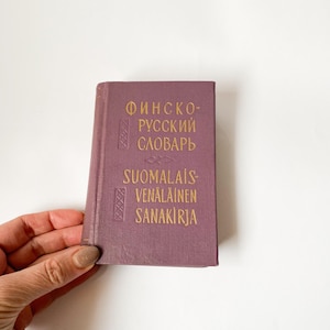 May include: A small, rectangular, purple book with gold lettering. The book's cover features text in both Cyrillic and Latin alphabets. The book is held by a hand with a neutral background.