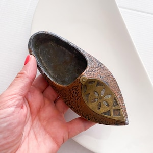 Puede incluir: Un pequeño cenicero en forma de zapato de color cobre con un diseño de latón en la punta. El cenicero tiene una textura martillada y un aspecto desgastado y antiguo.
