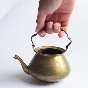 Pode incluir: Pequena chaleira antiga de latão com um bico curvo e uma alça decorativa. A chaleira tem um desenho floral gravado no corpo. A chaleira é segurada por uma mão contra um fundo branco.