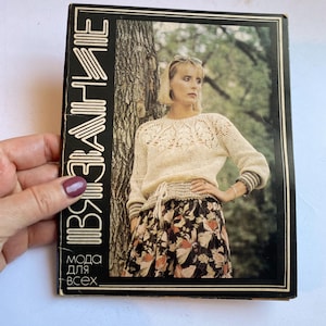 May include: A vintage book cover with the title in Russian. The cover features a woman in a cream-colored crocheted sweater and a floral skirt. The book is framed in black and held by a hand.