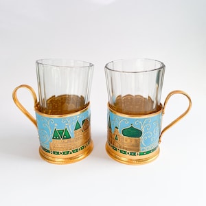 Könnte beinhalten: Zwei Teetassen aus Glas mit goldenen Griffen und dekorativen Haltern. Die Halter sind hellblau mit goldenen und grünen Akzenten und weisen ein Gebäudedesign auf. Die Tassen stehen auf einer weißen Oberfläche.