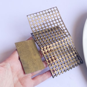 Op de afbeelding: Twee messing metalen stukken. Eén is een kleine rechthoek, de andere is een groter, geperforeerd vel met veel kleine, gelijkmatig verdeelde gaten. Het metaal heeft een warme, gouden tint en de randen van het geperforeerde vel zijn licht gebogen.