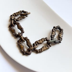 May include: A chain bracelet composed of interconnected bronze-toned metal links. Each individual link features a raised, ornate, scroll-like pattern with a darkened, weathered finish, creating a textured appearance throughout the entire piece.