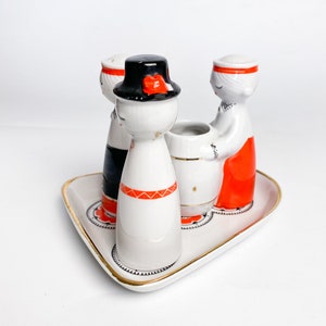 May include: A set of four porcelain salt and pepper shakers in the shape of people wearing traditional clothing. The shakers are arranged on a white triangular tray with a gold rim. The shakers are white with red and black accents.