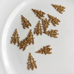 Könnte beinhalten: Eine Sammlung von zehn goldfarbenen Metall-Filigrananhängern, die jeweils eine längliche Dreiecksform mit komplizierten Schnörkeln, kleinen kreisförmigen Akzenten und mehreren Verbindungsösen an den Rändern für die Schmuckherstellung aufweisen.