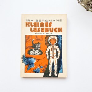 May include: A vintage book cover with the title "Kleines Lesebuch" in orange and black lettering. The cover features an illustration of a young boy in a white spacesuit with a red helmet and a cat in a spaceship.