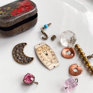 Puede incluir: Una colección de suministros y componentes vintage para la fabricación de joyas. Incluye una pequeña lata de metal oxidado, un dije de media luna de latón, una esfera de reloj, un cristal facetado y varias cuentas y hallazgos.