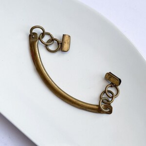 May include: A curved, antique brass-toned purse handle with attached rings and rectangular clasps. The handle is set against a white background, showcasing its vintage design and construction.