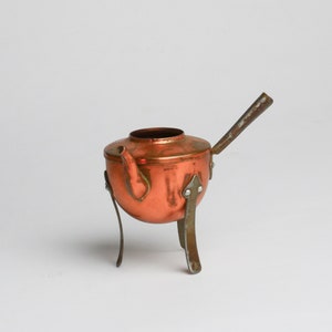 Op de afbeelding: Een koperen theepot met een handvat en drie poten. De theepot is gemaakt van koper en heeft een glanzende afwerking.