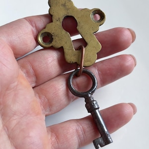 Könnte beinhalten: Ein Messing-Schlüsselschild mit dekorativem, gewelltem Rand und zwei Schraublöchern sowie einem zentralen Schlüsselloch. Ein kupferfarbener Metallring verbindet das Schild mit einem kleinen, dunkelgrauen antiken Metallschlüssel mit rundem Griff.