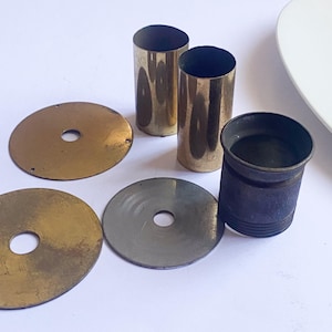 May include: A collection of vintage brass and metal components. The image shows two cylindrical brass tubes, two brass discs, one grey metal disc, and a dark blue metal cylinder. The components are arranged on a white surface.