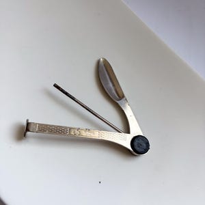 Könnte beinhalten: Ein silberfarbenes Pfeifenbesteck aus Metall mit drei klappbaren Werkzeugen: einem flachen Stopfer, einem dünnen Dorn und einem kleinen Löffel. Der Griff hat ein strukturiertes Rautenmuster und einen schwarzen runden Drehpunkt. Es ist geöffnet.