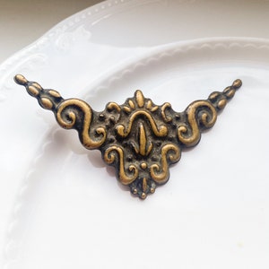 May include: A gold-toned metal decorative trim with a detailed ornate design. The trim has a curved shape with a central point and intricate scrollwork.